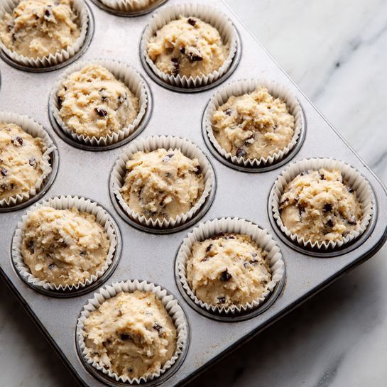 The image shows a close-up view of a metal muffin tray filled with white paper liners, each holding raw muffin batter. The batter is light beige with visible dark chocolate chips mixed throughout. The liners are neatly placed in the shallow cups of the tray, and the batter has a slightly rough texture with small lumps, filling each liner about three-quarters full. The background below the tray is a white marbled surface. photo taken with an iphone --ar 4:5 --v 7
