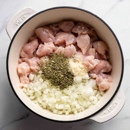 Inside a large white pot with a black rim, there are three layers visible: the bottom layer is finely chopped white onions that cover the pot's base; on top of the onions, there is a layer of raw small pink chicken pieces with a soft, smooth texture; on the very center, there is a small pile of green dried herbs mixed with light beige powdered seasoning, both resting on the chicken. The pot sits on a white marbled surface. photo taken with an iphone --ar 4:5 --v 7