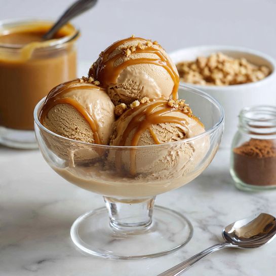 A clear glass bowl holds three round scoops of light brown ice cream with a smooth, creamy texture. Each scoop is topped with a thick drizzle of caramel sauce that glistens and runs down the sides. In the background, there is a white bowl filled with more caramel sauce, and a white bowl with granola or nuts on the right. A small glass jar with brown powder and a metal spoon lie on a white marbled surface. The setting is bright and softly lit, emphasizing the ice cream's rich texture and the shiny caramel sauce photo taken with an iphone --ar 4:5 --v 7