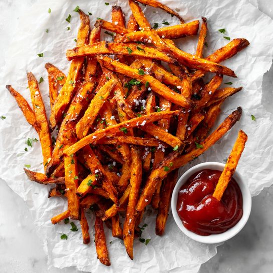 A large pile of long, thin sweet potato fries with a golden orange color and some darker brown edges lies loosely on crinkled white parchment paper that rests on a white marbled surface. The fries are sprinkled with coarse sea salt and small bits of green herbs. To the bottom right, there is a small white bowl filled with smooth, bright red ketchup, with two fries dipped into it, sticking out. The overall look is warm, crispy, and inviting. photo taken with an iphone --ar 4:5 --v 7