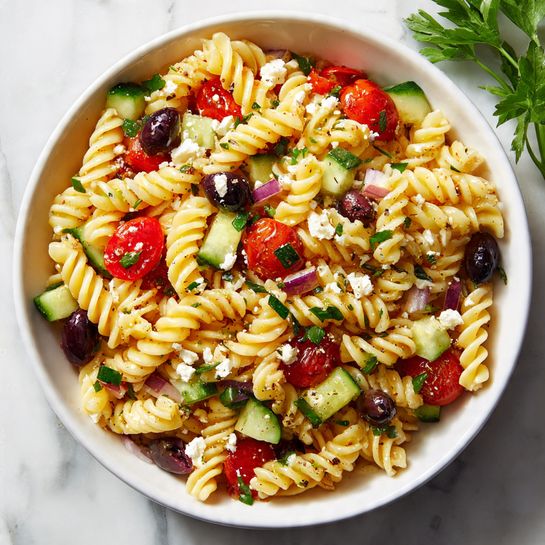 A white bowl filled with twisted rotini pasta as the base layer, pale yellow with a smooth texture. Mixed throughout are bright red cherry tomato halves and chunks of deep green cucumber, both scattered evenly. There are sliced black olives adding a dark contrast, with small pieces of white, crumbly feta cheese sprinkled on top. Bits of chopped parsley add flecks of vibrant green, along with small slivers of purple onion. The bowl rests on a white marbled surface with some scattered parsley leaves nearby. photo taken with an iphone --ar 4:5 --v 7