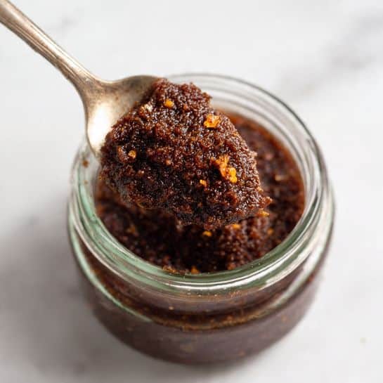 A close-up view of a glass jar filled with a dark brown, chunky textured paste that looks thick and slightly grainy with small visible bits throughout. Above the jar, a spoon holds a large scoop of this paste, showing its rich, dense consistency and a few orange specks mixed in. The jar and spoon are placed on a white marbled surface, creating a clean and simple background. The focus is sharp on the spoon and the mixture, while the jar's bottom part is blurred. photo taken with an iphone --ar 4:5 --v 7