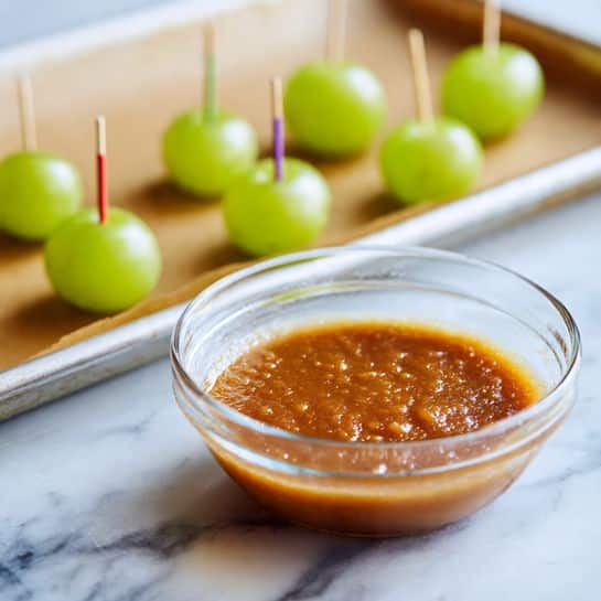 A clear glass bowl filled with a smooth, thick caramel-colored sauce with tiny lumps, placed on a white marbled surface. In the background, slightly out of focus, there is a baking tray lined with brown parchment paper. Seven bright green grapes are neatly lined up on the tray, each pierced by a colorful toothpick sticking straight up. The lighting is soft and natural, highlighting the glossy texture of the sauce. photo taken with an iphone --ar 4:5 --v 7