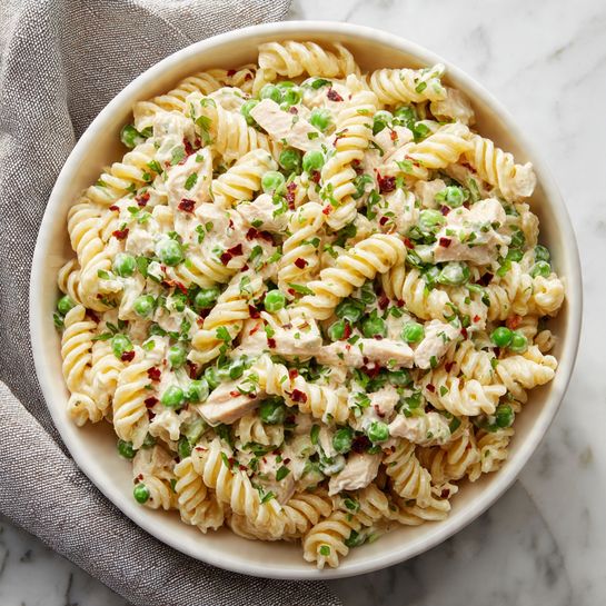 A large white bowl filled with three layers of creamy pasta salad. The bottom layer has small pale yellow pasta spirals mixed with small pieces of light beige chicken. The middle layer shows bright green peas scattered evenly throughout the pasta. The top layer is sprinkled with finely chopped green herbs and a few red flakes, adding a pop of color on the creamy white pasta. The bowl is placed on a white marbled surface with a gray cloth nearby. photo taken with an iphone --ar 4:5 --v 7