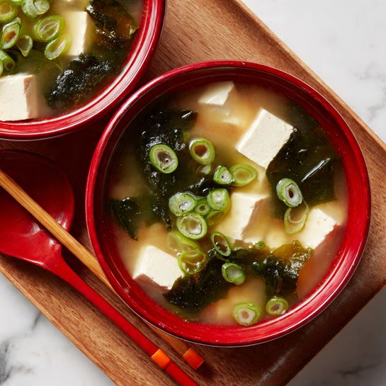 A close-up of a red bowl filled with light brown miso soup showing soft white tofu cubes scattered evenly in the broth. There are dark green pieces of seaweed floating across the liquid’s surface along with thin, ring-shaped slices of light green scallions spread around. The bowl sits on a light wooden tray with a pair of red and beige chopsticks resting on the tray’s side. The background is a white marbled texture. photo taken with an iphone --ar 4:5 --v 7