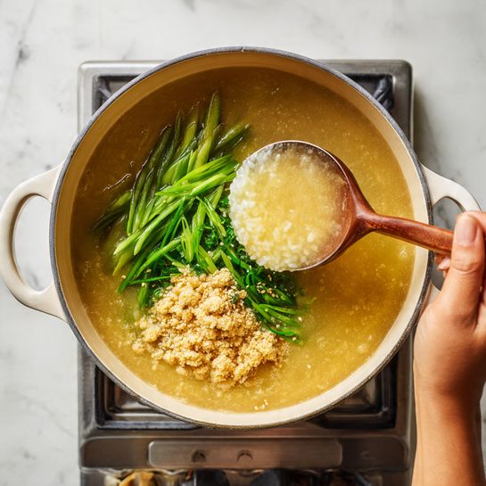 The dish shows a cooking pot with multiple layers inside. The first layer is cooked rice in a light brown broth filling most of the pot. A woman's hand pours more broth into the pot, creating a shiny, fluid surface. Next, a ladle holds thick rice and broth, showing a slightly creamy, soft texture. Then, green sliced scallions and light beige crushed nuts or seeds are added on top, floating in the broth. At last, a woman's hand stirs the mixture inside the pot with wooden chopsticks, mixing the green scallions and crushed nuts evenly into the creamy rice and broth. The background is a white marbled texture with a stove visible below the pot. Photo taken with an iphone --ar 4:5 --v 7