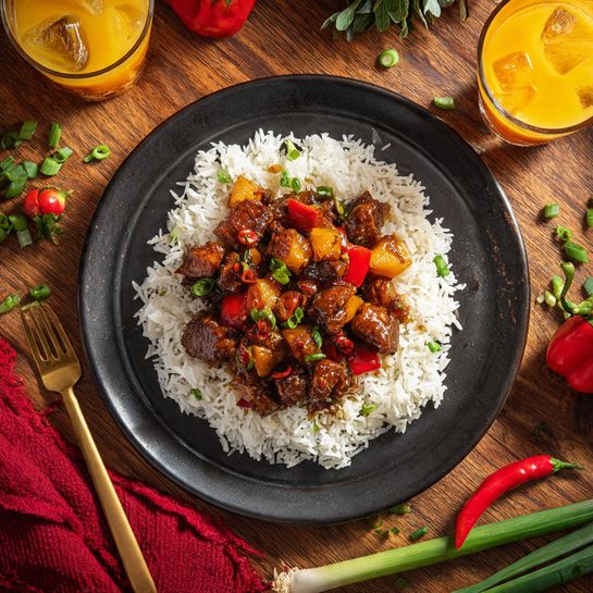 A black round plate holds a bed of white rice forming the base layer, topped with a thick brown stew containing pieces of meat and vegetables like potatoes and red peppers, all coated in a rich sauce with a slightly textured surface. The plate is placed on a wooden surface with scattered green onion pieces and whole green and red peppers around it. Two gold forks rest on opposite sides on the plate, and a glass of orange juice with ice cubes is seen in the top left corner. There are fresh herbs and a red cloth napkin near the bottom right of the plate. photo taken with an iphone --ar 4:5 --v 7
