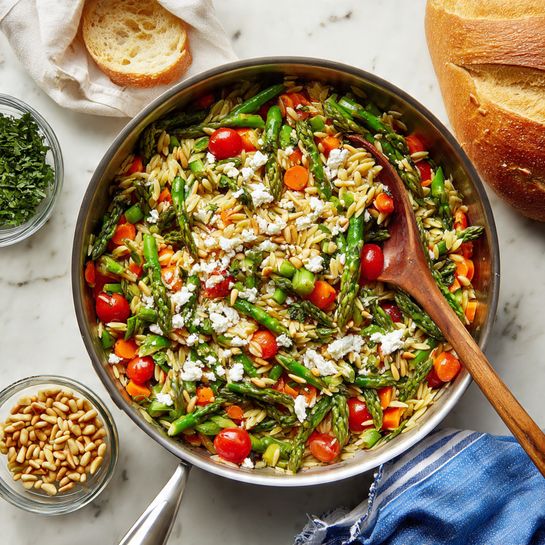 A large stainless steel pan filled with a colorful mix of sautéed vegetables, including bright green asparagus pieces, red cherry tomatoes, and small orange carrot chunks mixed with light brown orzo pasta. White crumbled cheese and toasted pine nuts are spread on top, adding texture and contrast. A wooden spoon is inside the pan, resting on the food. Around the pan, there is a small glass bowl of pine nuts, a small glass bowl of chopped green herbs, a round loaf of bread partially wrapped in white paper, and a blue-striped cloth napkin on a white marbled surface. Photo taken with an iphone --ar 4:5 --v 7