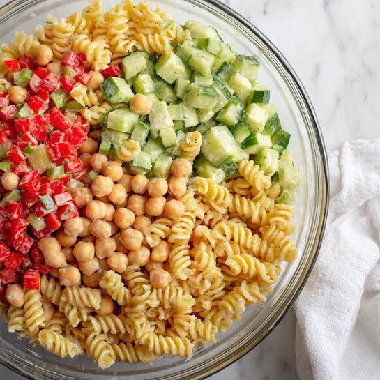 The image shows a close-up of a clear glass bowl filled with a pasta salad made of three visible layers. The bottom layer includes spiral pasta in a light yellow color mixed throughout. The middle layer has small chickpeas scattered evenly. The top layer shows a mix of diced green cucumber and small red bell pepper pieces distributed evenly, all coated in a creamy light dressing that slightly glistens on the ingredients. The bowl is set on a white marbled surface with some white fabric visible on the side. photo taken with an iphone --ar 4:5 --v 7