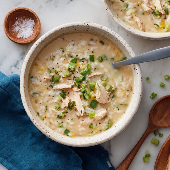 The image shows a bowl of thick rice soup with chicken and chopped green onions on top. The soup is creamy light brown with visible soft white chicken pieces mixed throughout the rice. The bowl is white with brown specks and has a gray soup spoon resting inside. Around the bowl, there is a small wooden bowl filled with coarse salt and a wooden spoon, scattered green onion pieces, and a blue cloth napkin on a white marbled surface. A part of another similar bowl of soup is visible in the upper right corner. Photo taken with an iphone --ar 4:5 --v 7