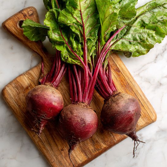 Three whole beets with deep red roots and long green leaves are placed on a wooden cutting board with a handle. The beets show some rough texture on their round roots, and their green leaves are fresh and slightly wrinkled, spreading outward at the top. The wooden board has a natural grain with light and dark brown shades and sits on a white marbled surface. photo taken with an iphone --ar 4:5 --v 7