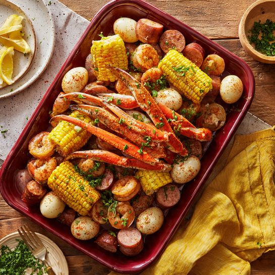 A deep red rectangular tray filled with a seafood boil showing bright orange crab legs and orange-pink shrimp scattered across the top layer, lightly dusted with spices and chopped green herbs. There are thick pieces of yellow corn on the cob and round white boiled eggs with some spice on their surface mixed in. Small golden-brown baby potatoes and slices of reddish-brown sausage form the base layer, with wedges of yellow lemon peeking through. The tray is placed on a rustic wooden table with a yellow cloth nearby and a white marbled textured background. photo taken with an iphone --ar 4:5 --v 7