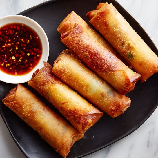 There are five crispy spring rolls with golden-brown and slightly shiny skins arranged in a neat row on a black square plate, showing some green and light fillings through the translucent wrap. A small white round bowl filled with a dark red chili dipping sauce, with visible chili flakes, is placed near the top left corner of the plate. The plate sits on a white marbled surface with soft natural light casting gentle shadows, highlighting the texture of the spring rolls and sauce. Photo taken with an iphone --ar 4:5 --v 7
