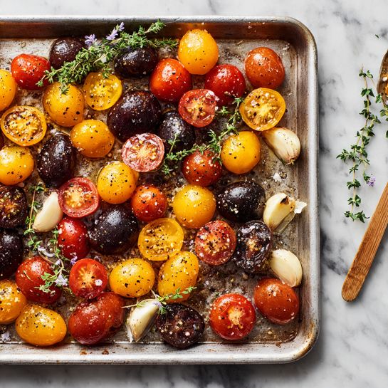 A baking tray with one layer of mixed small tomatoes in red, yellow, and dark purple shades scattered across it, some whole and some cut in half showing their juicy inside, sprinkled with black pepper and herbs. There are also several whole cloves of garlic and sprigs of green thyme with small purple flowers spread among the tomatoes. The metal tray appears used with brown roasted spots and seasoning marks. The scene is set on a white marbled surface. photo taken with an iphone --ar 4:5 --v 7