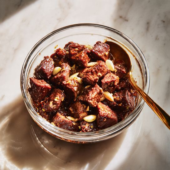 A clear glass bowl filled with chunks of dark brown marinated meat, each piece thick and coated with a shiny sauce. Small pieces of garlic are scattered among the meat, adding a touch of light beige color. A golden spoon rests inside the bowl on the right side, partially covered by the meat. The bowl is placed on a white marbled surface that softly reflects light. The scene is lit naturally from the left, creating warm highlights and soft shadows. Photo taken with an iphone --ar 4:5 --v 7