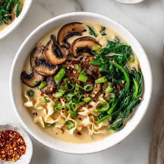 A white bowl filled with light beige broth and curvy, wavy, light cream noodles at the bottom layer. On top, dark green spinach leaves spread mostly over one side of the bowl. In the center, sliced brown mushrooms lay evenly, showing their pale inside and darker edges. Bright green chopped scallions are sprinkled over the mushrooms and spinach, mixed with small red chili flakes scattered around. The bowl is on a white marbled surface with a small white bowl of extra chili flakes nearby. photo taken with an iphone --ar 4:5 --v 7