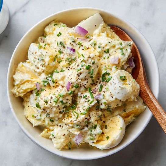A white bowl filled with creamy egg salad that has a pale yellow texture with white and yellow chunks of hard boiled eggs mixed in. The salad is topped with small chopped green herbs and tiny pieces of purple onion, with a light sprinkle of black pepper. A wooden spoon is placed inside the bowl, resting against the edge. The bowl is set on a white marbled surface with a blurred blue and white yogurt container in the background. photo taken with an iphone --ar 4:5 --v 7