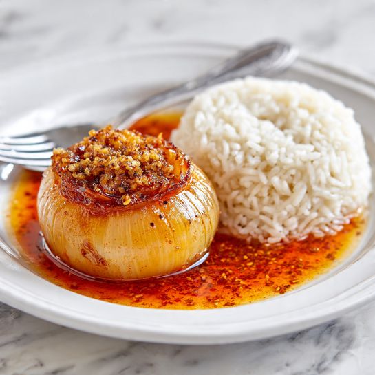 A white plate holds two main parts: on the right side, there is a large pile of white rice with soft, separate grains. On the left side sits a round, glazed onion with a golden-brown color and visible onion rings in the middle. Around the onion, an orange-red oily sauce with spices spreads slightly onto the plate, adding shine and texture. The plate is set on a white marbled surface, with a silver fork visible on the left edge. The overall look is clean and bright. photo taken with an iphone --ar 4:5 --v 7