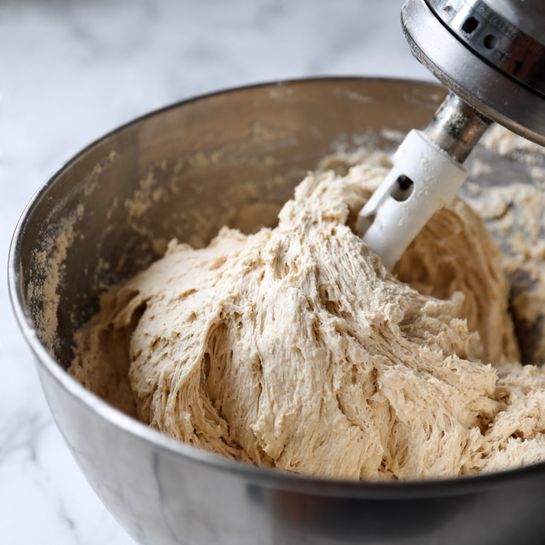 A large ball of light brown dough with a rough and slightly sticky texture is being mixed inside a silver metal bowl. The dough is attached to a white dough hook from a mixer, which is positioned above the dough and partially embedded in it. The dough appears thick and uneven, with folds and creases visible. The metal bowl has a slightly scratched surface and reflects light softly. The image is set on a white marbled surface. photo taken with an iphone --ar 4:5 --v 7