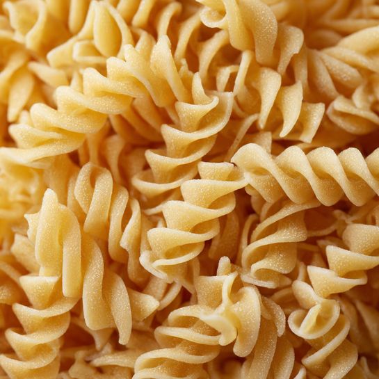 The image shows a close-up of many spiral-shaped pasta pieces, all pale yellow in color. Each pasta piece has a twisted, ridged texture, tightly packed and filling the whole frame. The lighting highlights the smooth but slightly rough surface of the dry pasta. The background is simple and clean, showing a white marbled texture. Photo taken with an iphone --ar 4:5 --v 7