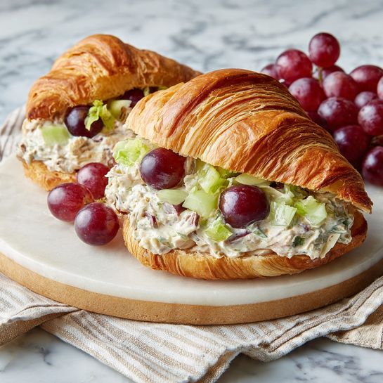 A golden brown croissant sliced in half horizontally holds a thick layer of creamy mixture filled with small chunks of light green celery and halved red grapes with dark purple centers. Behind the sandwich, a small bunch of red grapes is slightly out of focus. The croissant rests on a round white marble board with beige edging, placed over a beige and white striped cloth, all set against a white marbled surface. photo taken with an iphone --ar 4:5 --v 7