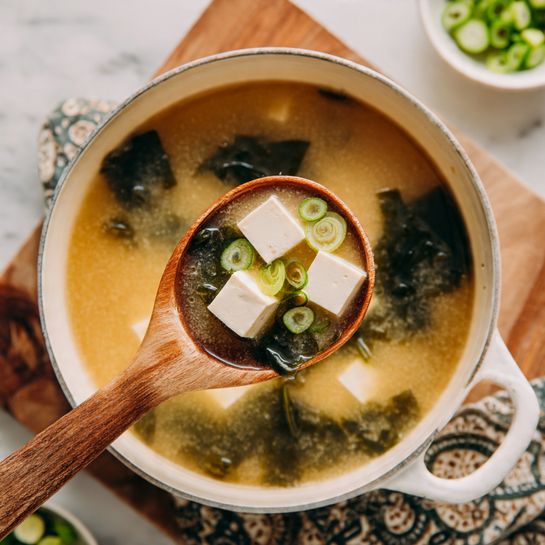 A close-up of a white saucepan filled with light brown miso soup that has smooth tofu cubes and dark green seaweed floating inside. A wooden spoon lifting some tofu cubes and small round green onion slices from the broth. The saucepan is on a white marbled textured wooden table with a patterned cloth under the handle. In the background, there is a small white bowl with sliced green onions. Photo taken with an iphone --ar 4:5 --v 7