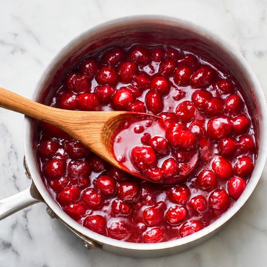 A silver metal pan sits on a white marbled surface, filled with a thick, glossy red fruit mixture that has visible chunks of fruit throughout. A wooden spoon with a smooth texture is resting inside the pan, partially covered in the red sauce. The sauce coats the sides of the pan unevenly, showing a rich and sticky texture. photo taken with an iphone --ar 4:5 --v 7