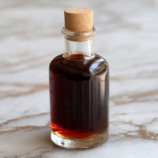 A clear glass bottle filled with dark brown liquid that has a slight shimmer inside. The bottle is cylindrical with a short narrow neck and is sealed with a light beige cork on top. The surface under the bottle has a white marbled texture with grey and black veins. The photo focuses closely on the bottle, showing its smooth reflective texture and the thick, rich liquid inside, with soft natural light highlighting its curves. photo taken with an iphone --ar 4:5 --v 7