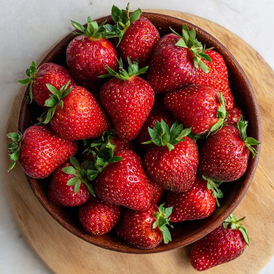 A round bowl filled with many bright red strawberries, each with small seeds and green leafy tops still attached. The strawberries are piled high, covering the entire bowl, which is on a light wooden surface. A few strawberries lie outside the bowl on the wood. The texture of the strawberries looks smooth and fresh, and the bowl itself is dark brown but the surface under it is a white marbled texture. photo taken with an iphone --ar 4:5 --v 7