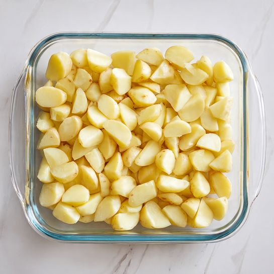 A clear glass baking dish filled with one layer of small potato pieces, each cut into uneven chunks showing both pale yellow skin and smooth light yellow inside flesh, all spread out evenly across the dish. The dish sits on a white marbled surface. photo taken with an iphone --ar 4:5 --v 7