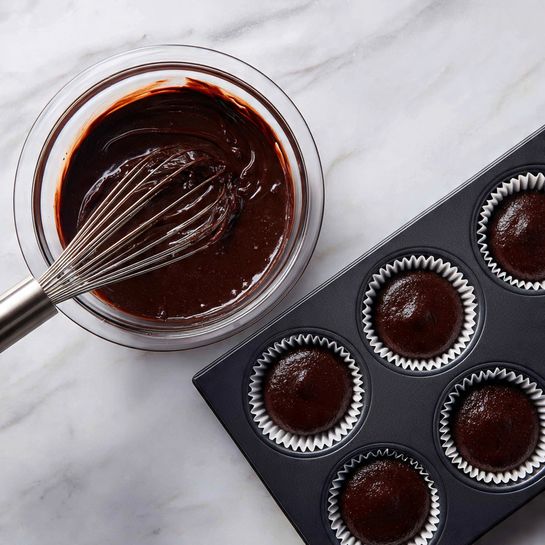 The image shows two parts. On the left side, there is a clear glass bowl filled with smooth, dark brown chocolate batter. A metal whisk is resting inside the bowl with some batter stuck on it, showing a shiny texture. The bowl sits on a white marbled surface. On the right side, a black muffin tray with six round sections is shown. Each section has a white paper liner filled with the same dark brown chocolate batter, smooth and shiny on top, waiting to be baked. The tray is also on a white marbled surface. photo taken with an iphone --ar 4:5 --v 7