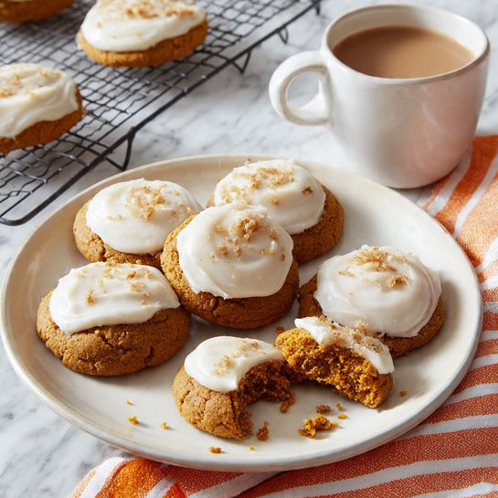 The image shows a white round plate with seven pumpkin cookies topped with a thick layer of smooth white frosting arranged on it. One cookie is broken into two pieces with crumbs scattered nearby. Next to the plate is a white cup filled with light brown hot chocolate or coffee, sitting on a white and orange-striped cloth. In the top left corner, there is a cooling rack with a single pumpkin cookie on it. Everything is placed on a white marbled surface. photo taken with an iphone --ar 4:5 --v 7