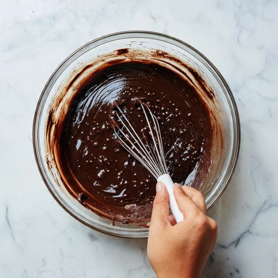 A clear glass bowl filled with dark brown, glossy chocolate batter that has a smooth and thick texture, with small bubbles visible on the surface. A woman's hand is seen holding a white-handled whisk, mixing the batter in the bowl. The bowl sits on a white marbled surface, with some chocolate batter smeared slightly on the sides of the bowl. photo taken with an iphone --ar 4:5 --v 7