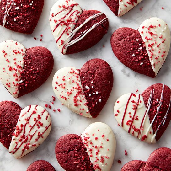 Several heart-shaped cookies are shown, made of bright red dough. Each cookie has white chocolate drizzled on top in different ways: some have thick strips covering half of the cookie, others have thin lines spread across. Red sugar sprinkles cover part of the white chocolate layer on some cookies. The cookies are arranged on a round white plate with a smooth texture, which is placed on a white marbled surface. To the right, there is a small clear glass bowl filled with red sugar sprinkles. In the bottom left corner, there is a small piece of white chocolate bar. Photo taken with an iphone --ar 4:5 --v 7