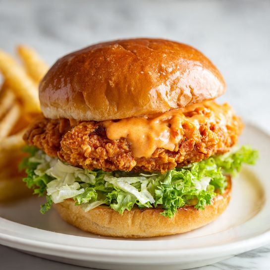 A crispy golden brown fried chicken patty sits on a bed of shredded light green lettuce, all inside a soft, shiny, golden brown bun with a smooth glossy top. A light pink creamy sauce drips slightly from the crispy chicken. The sandwich rests on a dark gray plate, placed on a white marbled surface with a soft textured cloth underneath. In the background, there is a small bowl with sliced red tomatoes and a small container of sauce, both slightly blurred. photo taken with an iphone --ar 4:5 --v 7