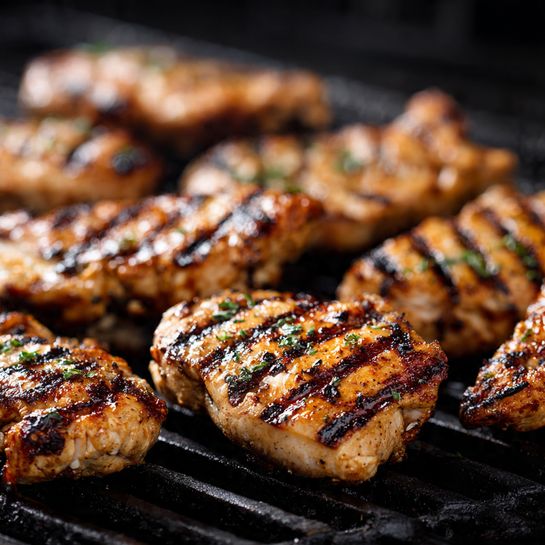 Many pieces of grilled chicken lie close to each other on a black grill. Each piece is thick and has clear dark grill marks running diagonally across its light brown, slightly shiny surface. The texture of the chicken looks moist with some charred spots adding a darker brown color. The pieces vary slightly in size but all share the same cooked look. The background is blurred black grill lines under the chicken. photo taken with an iphone --ar 4:5 --v 7