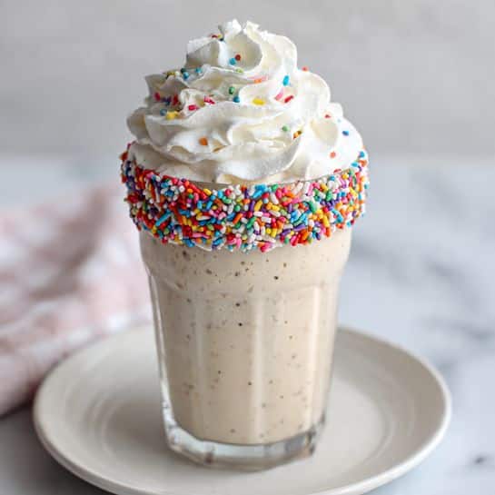 A tall clear glass filled with a light beige milkshake that has small vanilla bean specks visible inside, topped with a large swirl of white whipped cream. The glass rim is coated with a thick layer of multicolored round sprinkles that add vibrant colors around the top edge. The glass sits on a white plate placed on a white marbled surface with a soft, blurred white background. Photo taken with an iphone --ar 4:5 --v 7