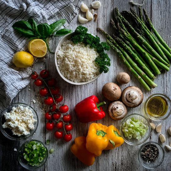 The image shows fresh, colorful ingredients neatly arranged on a dark wooden surface with a white marbled look subtly edited in the background. On the left, there is a striped grey cloth with a white bowl filled with raw white rice topped with a bunch of green parsley. Next to it, there are long green asparagus spears and clusters of small red cherry tomatoes on the vine. Two lemon halves lie near the asparagus. At the bottom left, a clear bowl contains crumbled white cheese, and another small group of cherry tomatoes sits nearby. Moving rightwards, there are several small colorful bell peppers – red, yellow, and orange – placed close together. Three whole brown mushrooms, some peeled garlic cloves, and sliced shallot with ginger are next to a small glass bowl filled with sliced green onions. A clear bowl of golden olive oil and another with salt and pepper complete the setup. Bright green basil leaves add a fresh touch. Everything looks clean and fresh, captured clearly and brightly. photo taken with an iphone --ar 4:5 --v 7