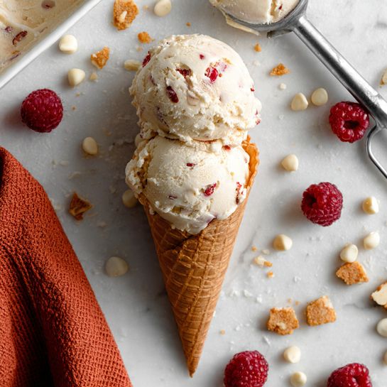 A waffle cone holds two scoops of creamy white ice cream with visible small pieces of red raspberries mixed inside, showing a rough texture. Around the cone, fresh whole red raspberries and small white chocolate chips are scattered on a white marbled surface, along with broken pieces of a golden brown cookie or cracker. A metal ice cream scoop with some ice cream rests on the surface near the cone, and a corner of a white container filled with the same raspberry ice cream is partially visible. An orange textured cloth is placed to the left side of the image. photo taken with an iphone --ar 4:5 --v 7
