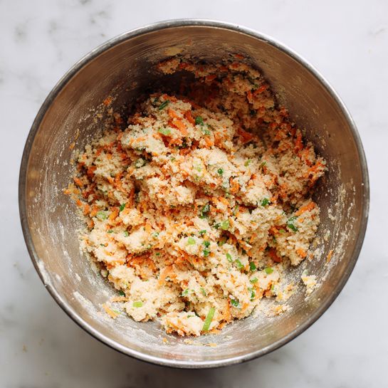A large metal bowl filled with a thick, crumbly mixture that has a pale beige base with small pieces of bright orange carrot and green spring onion scattered evenly throughout, creating a rough texture and a colorful contrast. The mixture looks slightly dry but with some moisture, and the inside of the bowl shows some residue from mixing. The image sits on a white marbled surface. photo taken with an iphone --ar 4:5 --v 7