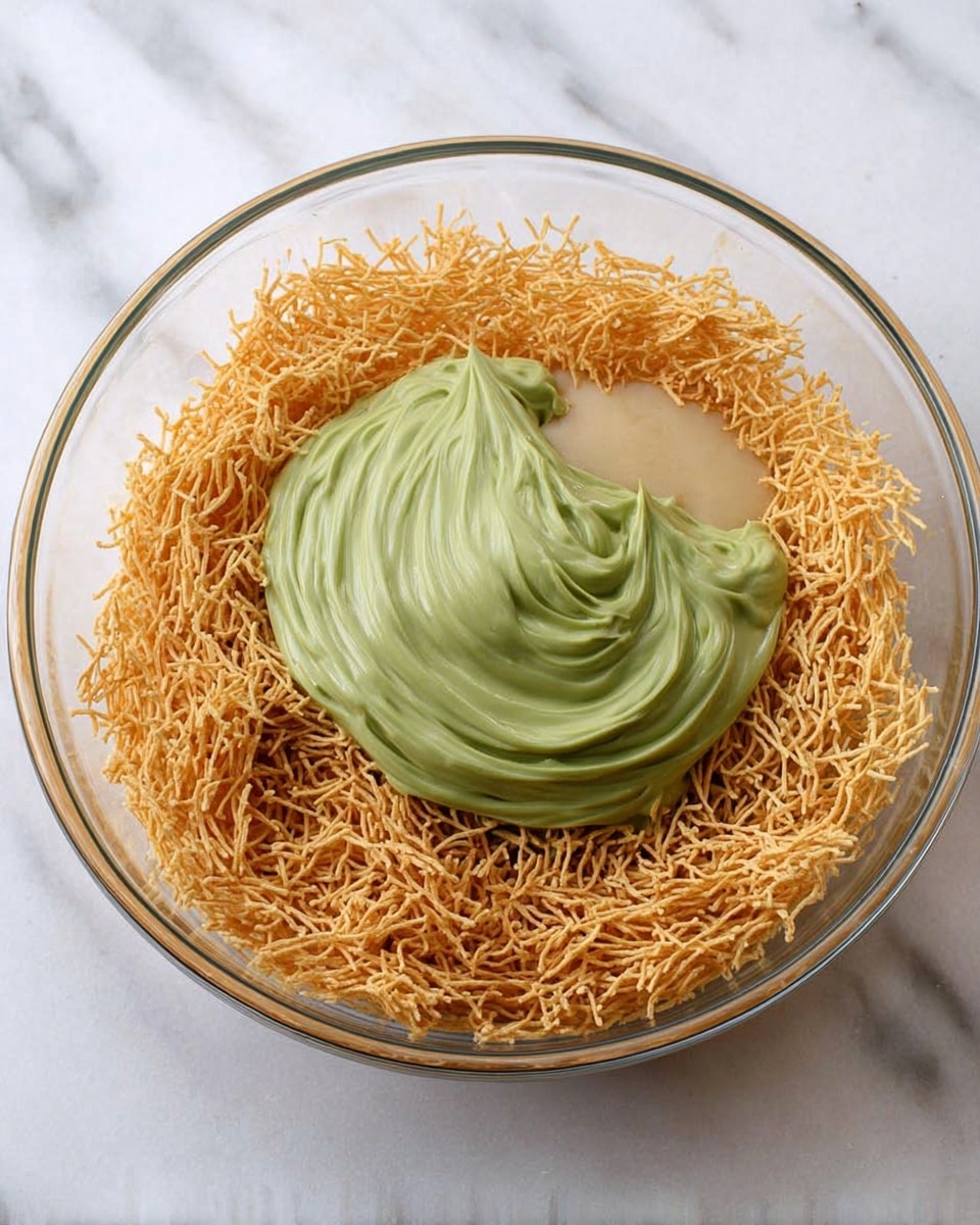 The image shows a clear glass bowl filled with three layers of ingredients. The bottom layer is made of thin, light golden-brown crispy strands spread evenly inside the bowl. On top of this is a thick, creamy green mixture placed in the center with smooth swirled texture. Next to the green mixture, there is a light beige sauce or liquid filling a small section within the crispy strands. The bowl sits on a white marbled surface. photo taken with an iphone --ar 4:5 --v 7