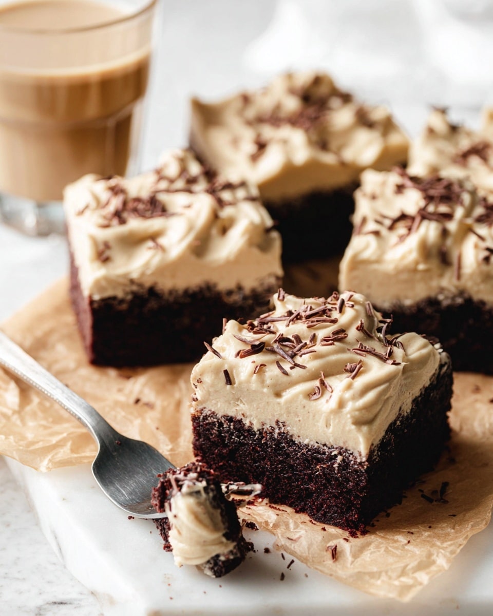 The image shows four square chocolate cake pieces placed on brown parchment paper on a white marbled surface. Each cake piece has two layers: a dark brown, moist-looking cake base topped with a thick, creamy light beige frosting. The frosting is textured with soft swirls and curls, and each piece is sprinkled with small, thin chocolate shavings. One cake piece is in the front with a silver fork partially stuck into its side, lifting a small bite out. In the background, slightly blurred, is a clear glass with a light brown creamy drink. Photo taken with an iphone --ar 4:5 --v 7