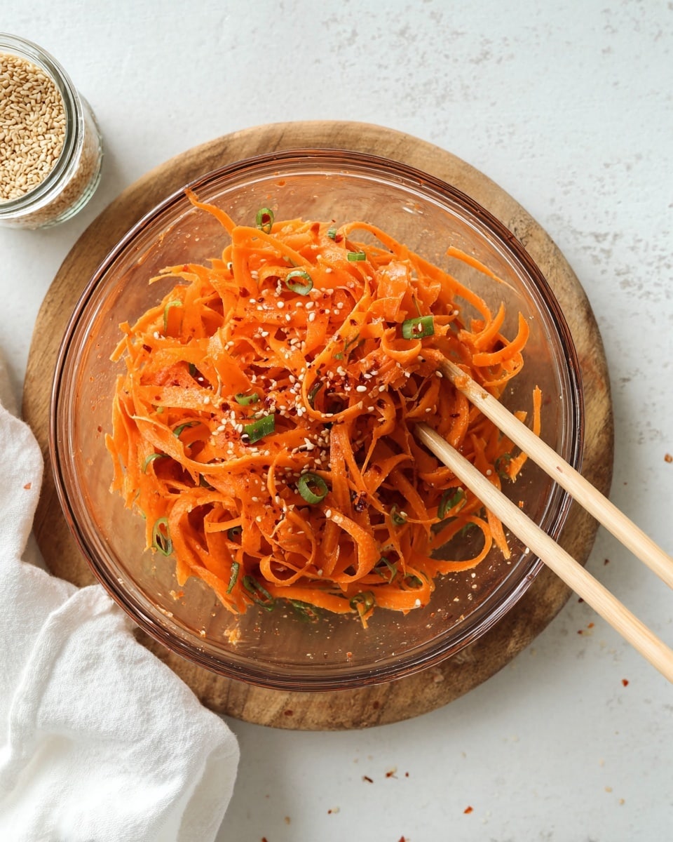 A clear glass bowl sits on a round wooden board, filled with bright orange carrot ribbons that are thin and curly, mixed with small green slices of scallion and sprinkled with white sesame seeds and red chili flakes. The carrot ribbons have a wet, glossy look from the dressing, and two light wood chopsticks rest in the bowl, slightly lifting some carrot ribbons. The bowl is placed on a white marbled surface with a white cloth napkin nearby, and an open jar of sesame seeds is lying on its side close to the bowl. photo taken with an iphone --ar 4:5 --v 7