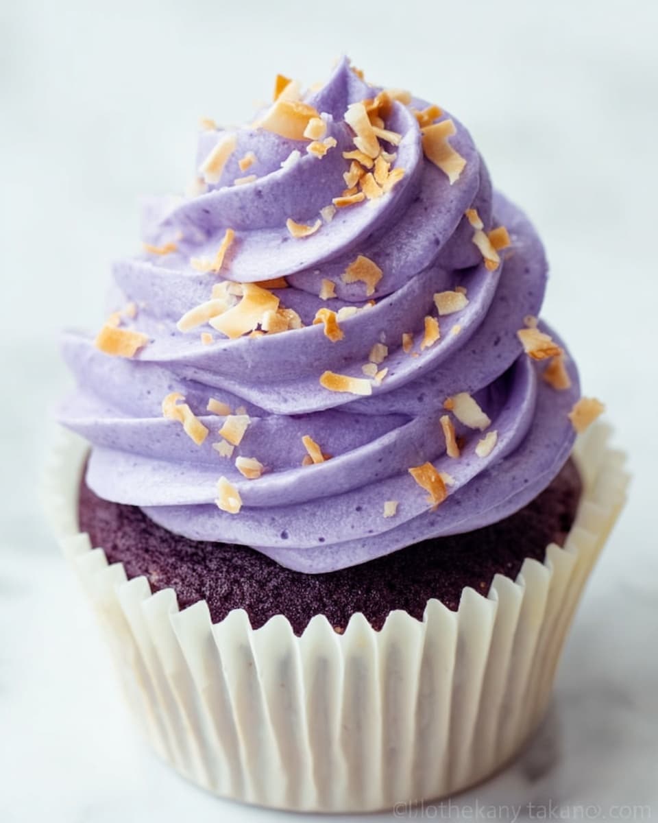 A white cupcake liner holds a rich dark purple cupcake base. On top, there are three thick swirls of light purple, creamy frosting stacked high, each layer smooth and airy in texture. Small golden toasted coconut flakes are scattered all over the frosting, adding a crunchy texture contrast. The background is a white marbled texture, soft and clean. photo taken with an iphone --ar 4:5 --v 7