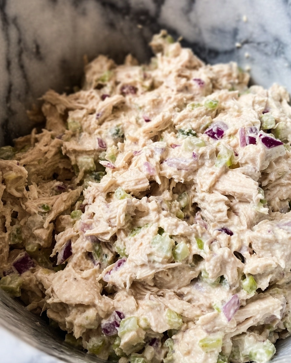 The image shows a close-up of a creamy chicken salad mixture with about three layers of texture visible: shredded light beige chicken pieces, small chunks of white and purple onion, and finely chopped green celery. The ingredients are mixed thoroughly, creating a mostly light beige color with specks of white, green, and purple scattered throughout. The mixture looks moist and slightly chunky, with a soft, creamy dressy texture. It is inside a container with a white marbled texture background. photo taken with an iphone --ar 4:5 --v 7