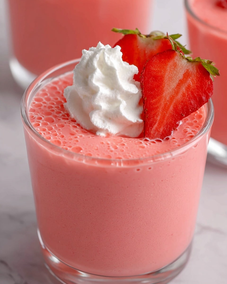 A glass filled with a smooth, bright pink strawberry mousse with small air bubbles visible on the surface. On top, there is a swirl of white whipped cream placed slightly off-center, and two fresh strawberry slices—one whole and one cut—are arranged next to the cream, showing their bright red color and seeds clearly. The glass sits on a white marbled surface, with another similar glass blurred in the background. photo taken with an iphone --ar 4:5 --v 7