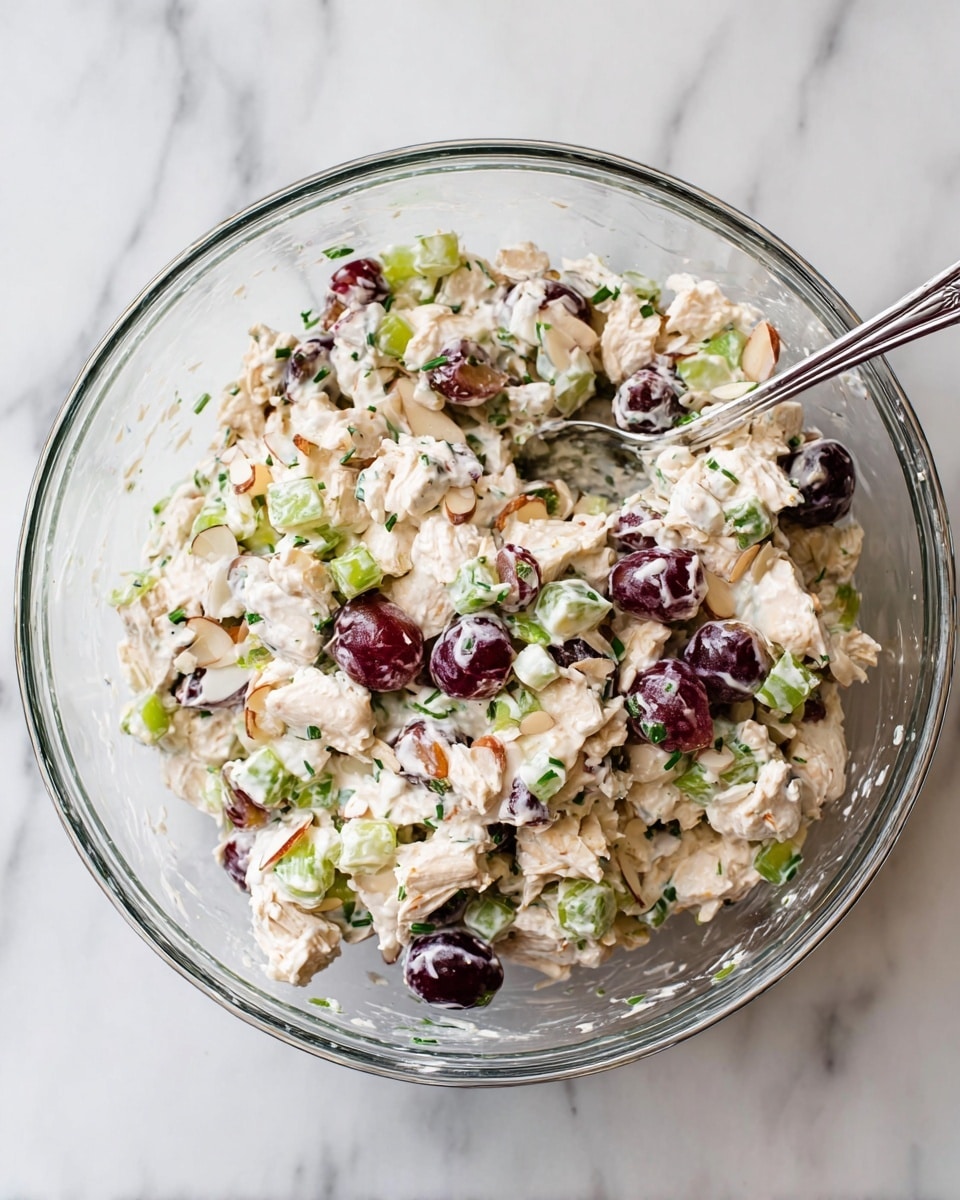 A clear glass bowl is filled with a creamy chicken salad mixture. The salad has three main layers visible throughout: off-white creamy dressing mixed with chopped light beige chicken pieces, deep red and purple grape halves, and bright green chopped celery and green onions. There are also thin almond slices mixed in for texture. A long silver spoon is resting inside the bowl. The bowl sits on a white marbled surface, with some small bits of herbs scattered inside the bowl. photo taken with an iphone --ar 4:5 --v 7