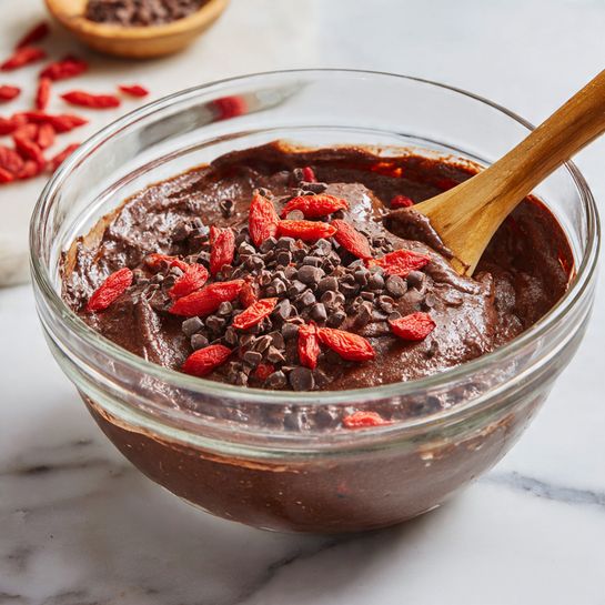 A clear glass bowl contains dark brown thick chocolate batter with a smooth and rich texture. On top of the batter, there are small clusters of dark brown cacao nibs and bright red dried goji berries, adding contrasting colors and rough textures. A wooden spoon with a light brown handle rests inside the bowl, partly submerged in the batter, with part of its handle leaning against the bowl's side. The bowl sits on a white marbled surface with blurred bits of red and brown ingredients in the background. Photo taken with an iphone --ar 4:5 --v 7