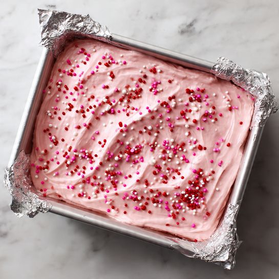 The image shows a single thick layer of smooth, light pink frosting spread evenly inside a foil-lined white rectangular pan. The frosting has soft, round swirls and small ripples all over its shiny surface. Scattered across the frosting are many tiny round sprinkles in dark red, bright pink, white, and light pink colors, adding a speckled pattern. The pan rests on a white marbled surface. photo taken with an iphone --ar 4:5 --v 7