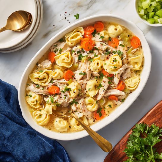 A white oval dish filled with a soup made of three layers: the bottom layer is clear light brown broth, the middle layer is small yellow tortellini pasta, and the top layer has shredded light brown chicken pieces, bright orange carrot chunks, and green celery slices, all mixed together with small black pepper specks visible. A gold spoon rests inside the dish, scooping some pasta, chicken, carrots, and celery. The dish is placed on a white marbled surface with a navy blue cloth nearby, a white stack of bowls with golden spoons, chopped celery on a wooden board, and some green parsley sprigs scattered around. Photo taken with an iphone --ar 4:5 --v 7